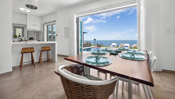 A modern kitchen with white cabinets, a dining area with a wooden table and chairs, and a large window offering a view of the sea.