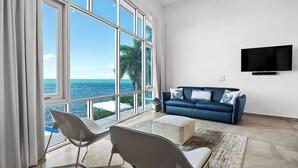 A modern living room with a large window overlooking the sea, a blue sofa, and a flat-screen TV on the wall.