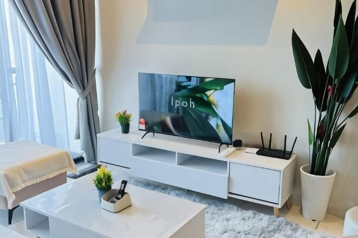 A modern living room with a flat-screen TV, white furniture, a potted plant, and a window with curtains.