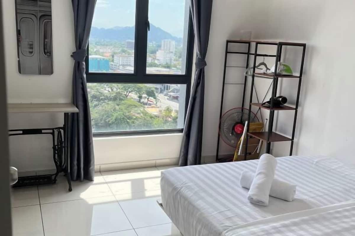 A hotel room with a bed, a standing fan, a shelf with items, and a window with a view of buildings and trees.