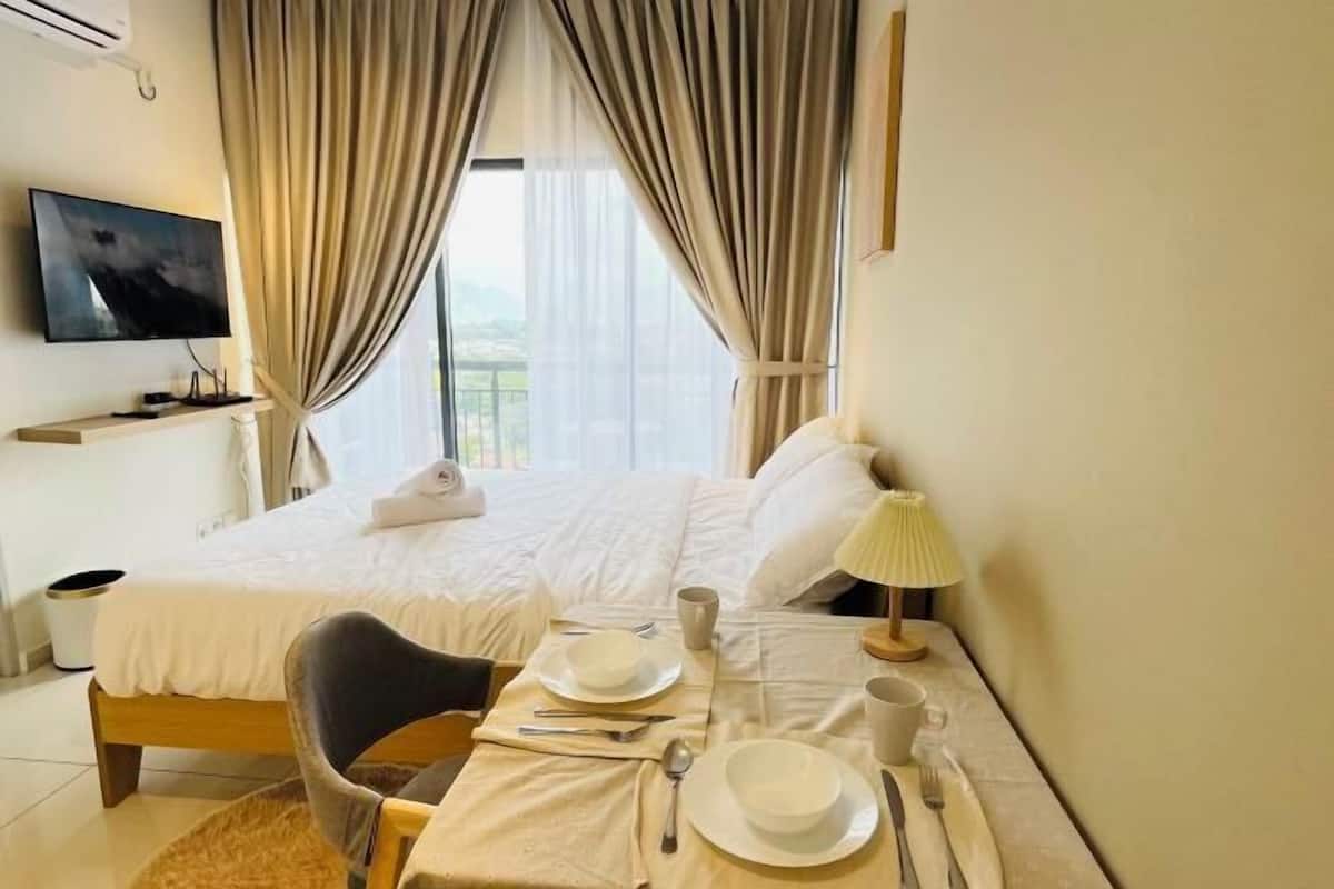 A hotel room with a bed, a TV mounted on the wall, a dining table with plates, cups, and cutlery, and a lamp.