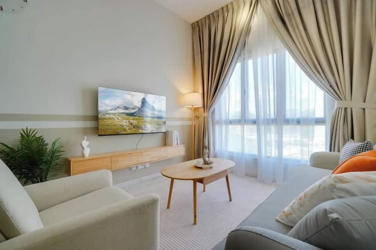 A modern living room with a sofa, a coffee table, a TV mounted on the wall, and a large window with curtains.