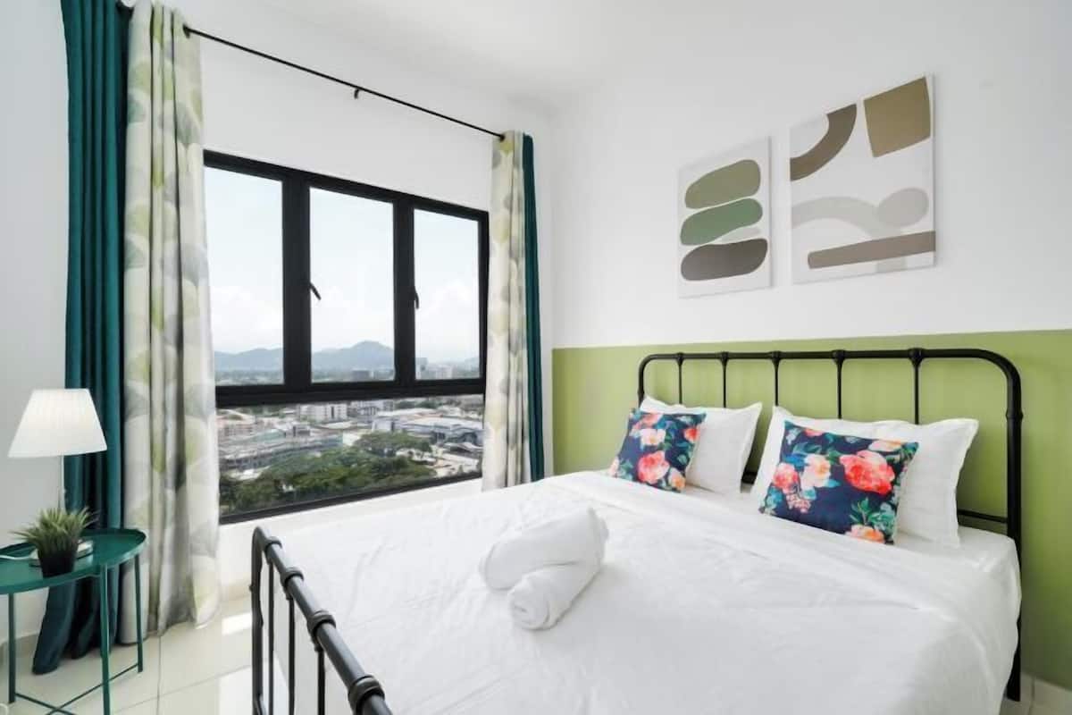 A hotel room with a large window, a bed with floral pillows, a green headboard, and a small table with a lamp.