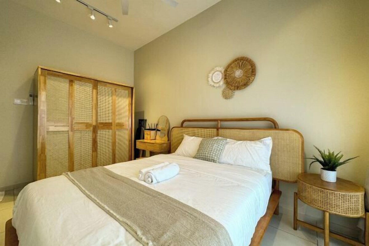 A neatly made bed with a white comforter and a rolled towel, a wooden headboard, a small bedside table with a plant, and a woven screen door.
