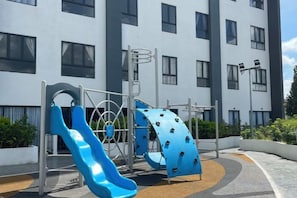 A playground with slides and climbing structures, located in front of a modern building with multiple windows.