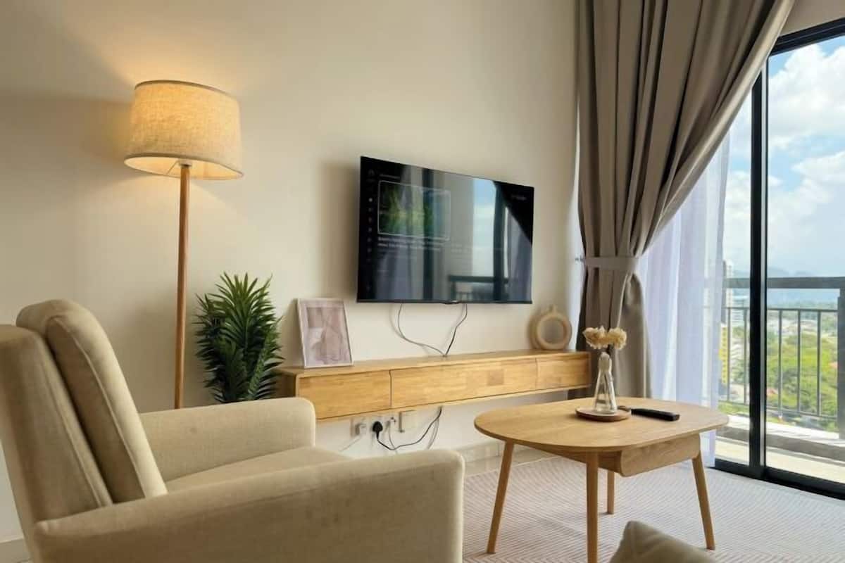 A modern hotel room with a sofa, a round wooden table, a TV mounted on the wall, a potted plant, and a view of the outdoors through large windows.
