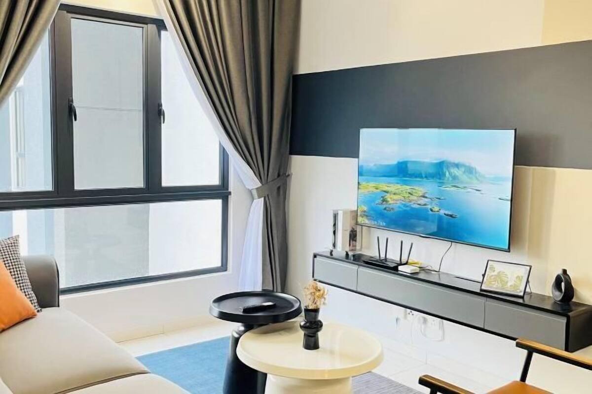 A modern living room with a large flat-screen TV, a sofa, a round coffee table, and a view of a body of water through a window.