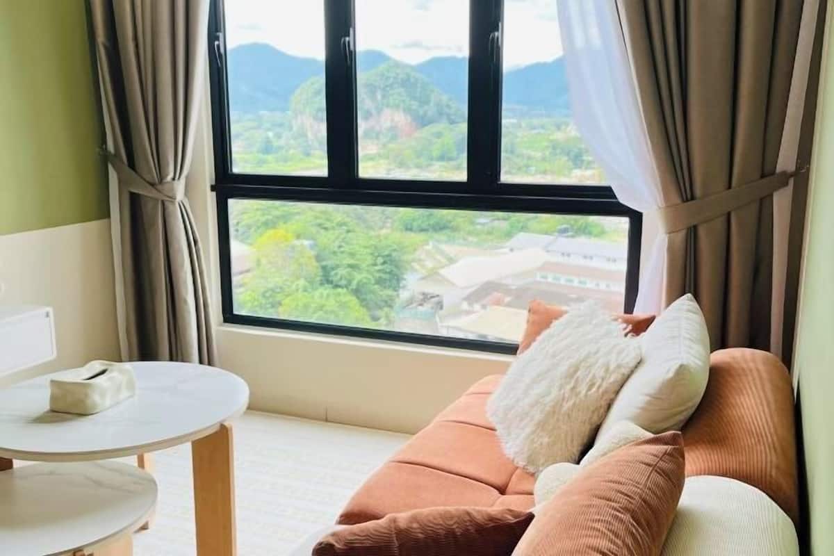 A room with a sofa, a table, and a window with a view of greenery and buildings.
