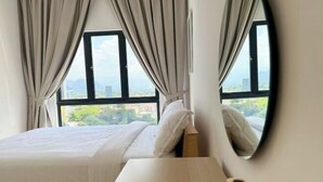 A hotel room with a bed, a desk, and a view of a cityscape through large windows.