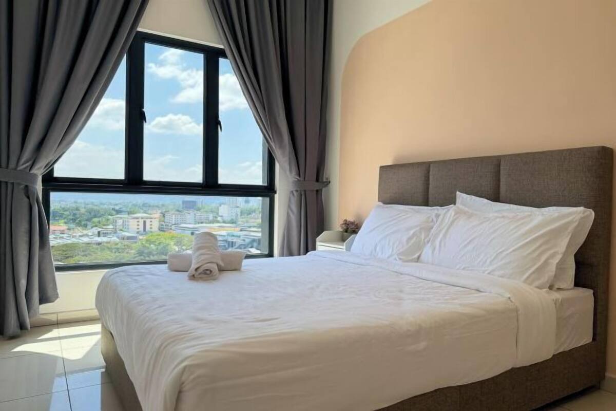 A neatly made bed with white linens and pillows, a grey headboard, and a view of a cityscape through a large window with grey curtains.