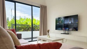 A modern living room with a large flat-screen TV mounted on the wall, a sofa with red cushions, a white coffee table, and a balcony with green plants.