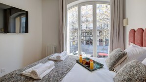 Ein Hotelzimmer mit großem Fenster, einem Bett mit Frühstücksbrettchen, einem Heizkörper und einem Spiegel an der Wand.