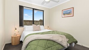 Une chambre à coucher avec un lit, une fenêtre, un ventilateur de plafond, une table de chevet avec une lampe et un tableau encadré au mur.