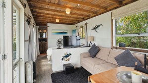 A cozy room with a wooden ceiling, a sofa, a TV, and a window with a view of greenery. - Tawharanui Cabin - Rural Cabin Studio (Warkworth)
