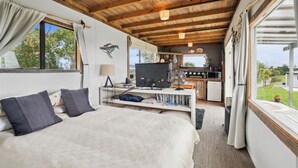 A spacious bedroom with a large bed, a desk with a computer, and a view of a lush garden. - Tawharanui Cabin - Rural Cabin Studio (Warkworth)