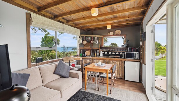 A cozy living area with a sofa, a dining table, and a kitchen area with a microwave and a refrigerator. - Tawharanui Cabin - Rural Cabin Studio (Warkworth)