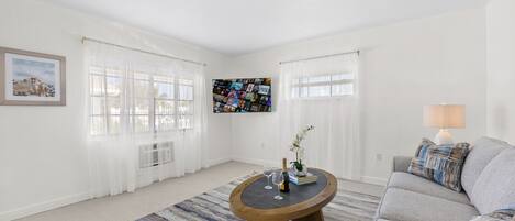 Una sala de estar con un sofá gris, una mesita redonda con un florero y dos vasos, un cuadro enmarcado en la pared y un gran collage de fotografías.