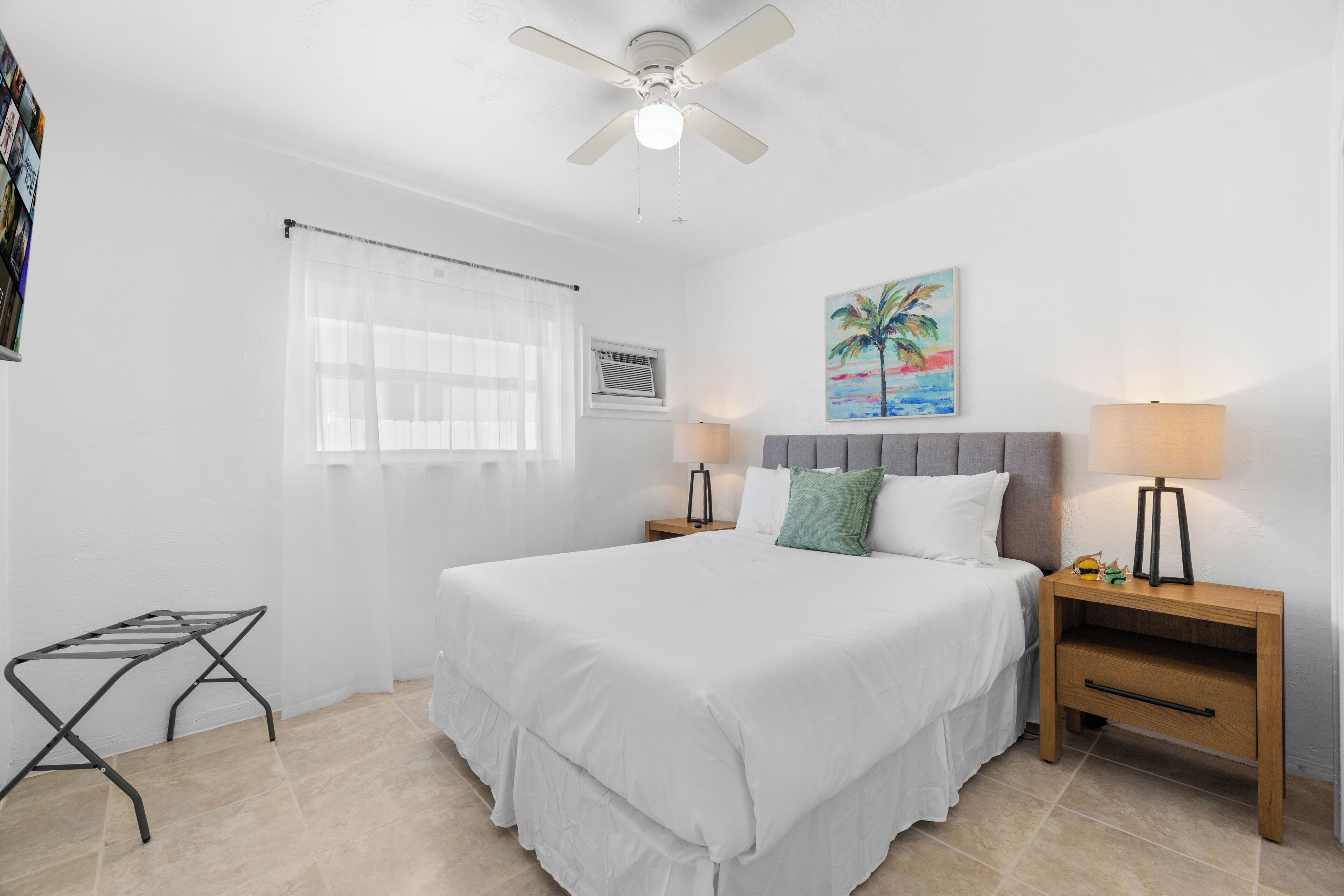 A neatly arranged bedroom with a bed, two bedside tables with lamps, a ceiling fan, a window with sheer curtains, and a wall-mounted air conditioning unit.