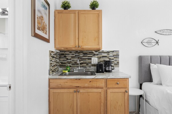 A compact kitchen with wooden cabinets, a tiled backsplash, and a small countertop. There is a sink, a coffee maker, and a plant on the counter.