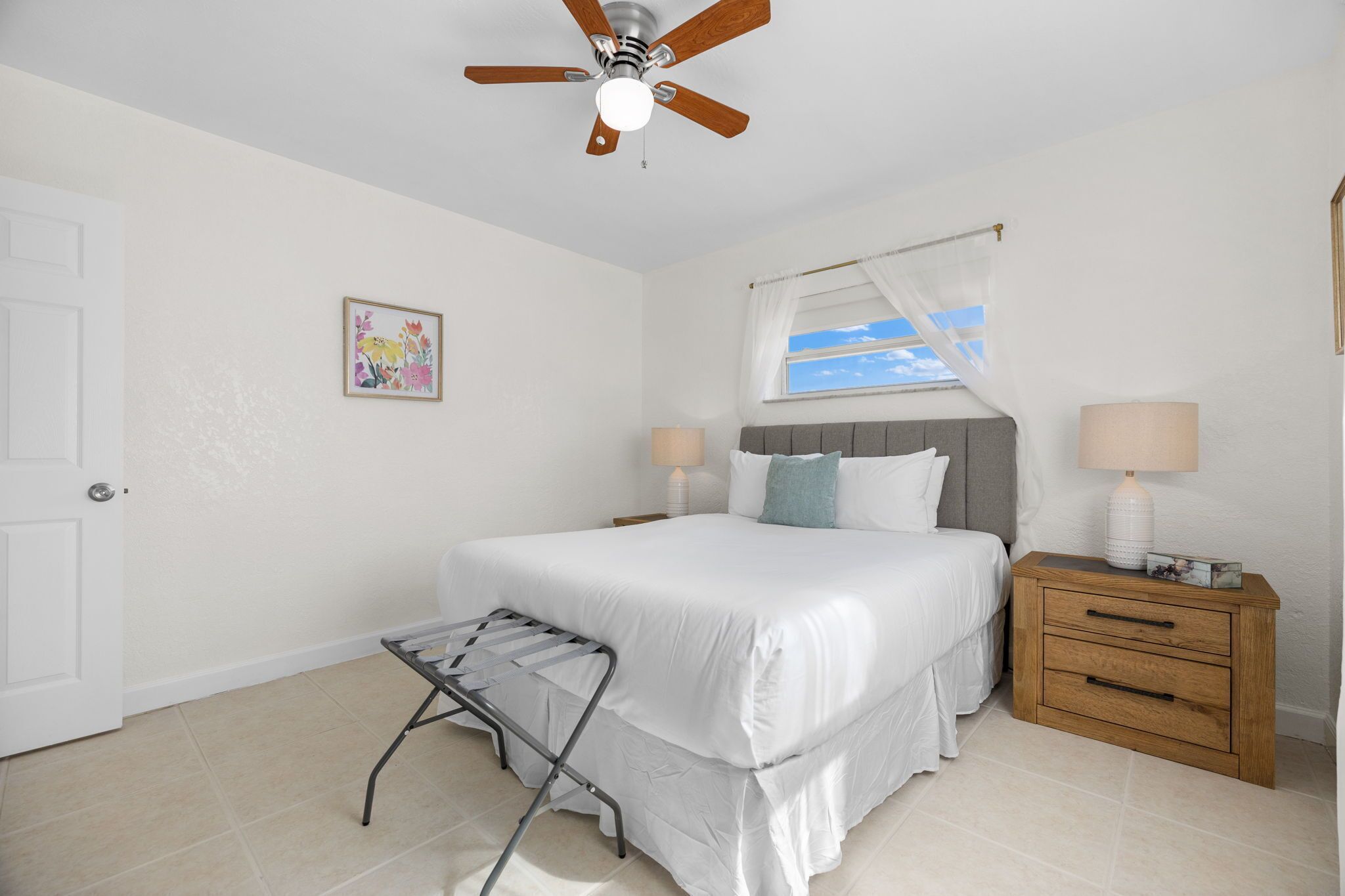 A bedroom with a bed, a ceiling fan, a window with a view, a bedside table with a lamp, and a small stool.