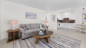 Un salon moderne avec un canapé gris, une table basse en bois et un tapis à rayures. Au fond, on aperçoit une cuisine avec des meubles blancs et un coin repas comprenant une table et des chaises.