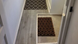 A narrow hallway with a wooden floor, a patterned rug at the entrance, and a door leading to a room with a bed and a chair.