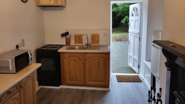 A compact kitchen with wooden cabinets, a built-in microwave, a sink, and a stove. There's a clock on the wall and a door leading outside.