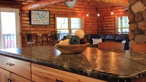 Interior de una cabaña de madera con pared de piedra, una mesa de comedor de madera, un sofá y un cuenco con adornos en la encimera.