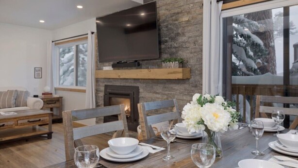 A dining area with a wooden table set for four, a fireplace, and a television mounted on a stone wall. - Walkable to Canyon Lodge w/ Ski Locker (Mammoth Lakes)