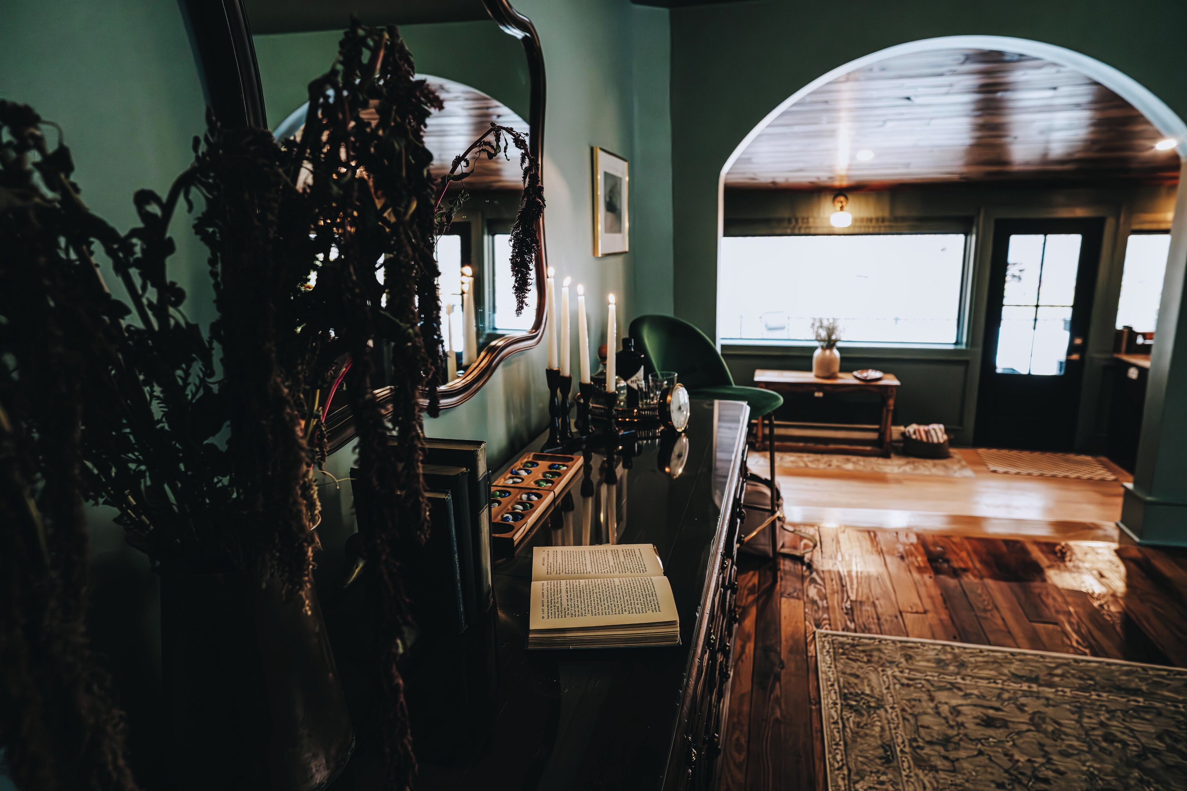 Habitación con un espejo grande, un libro y una silla verde. Se observan ventanas con cortinas y techo de madera.