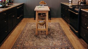 A kitchen with dark wooden cabinets, a wooden table, a rug, and a hanging light fixture.