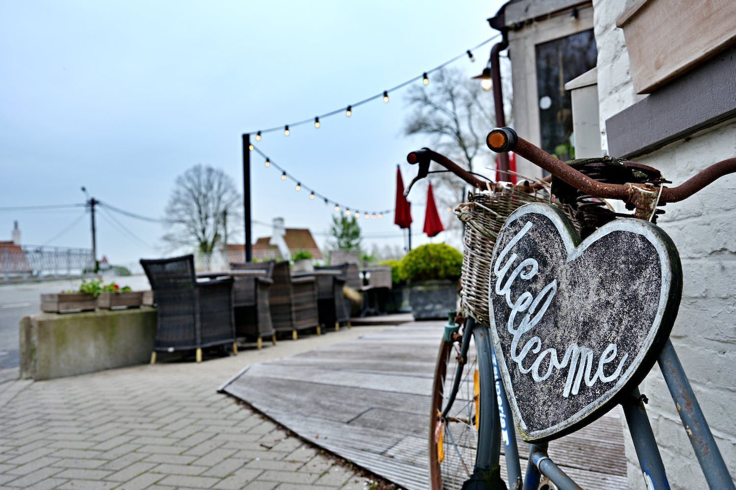 A bicycle with a heart-shaped sign that reads "Welcome" attached to it, parked on a paved path with outdoor seating and string lights in the background.