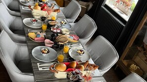 Una larga mesa de comedor dispuesta con alimentos para el desayuno, como pan, bollería y fruta, en una habitación moderna con sillas grises y vistas al exterior.