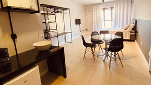 Une chambre d’hôtel compacte comprenant un coin cuisine, un espace repas avec une table et des chaises, un lit avec un téléviseur et une fenêtre avec des rideaux.