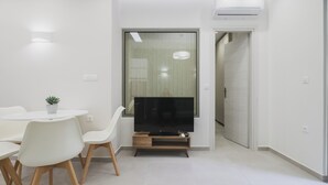 Ein modernes Wohnzimmer mit einem Flachbildfernseher auf einem Holzständer, weißen Sesseln, einem kleinen Topfpflanzen und einer Schiebetür zu einem anderen Raum.