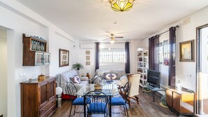 Een ruime woonkamer met een glazen salontafel, blauwe fauteuils, een houten kast en een plafondventilator.