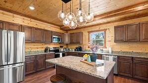 A modern kitchen with wooden cabinetry, stainless steel appliances, granite countertops, and a central island with stools. - Brand New Greenleaf Lodge ~Indoor Pool ~ Hot Tub (Sevierville)