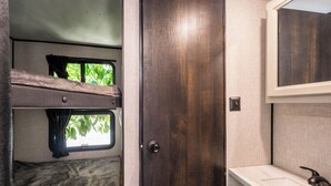 A compact RV interior with a bunk bed, a small window with curtains, a sink, and a mirror. - The Happy Camper - Cozy RV sleeps 4, with WiFi, in beautiful Ojai (Ojai)