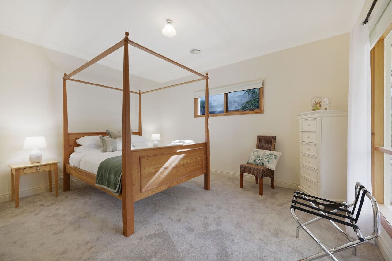 Uma cama com dossel em quarto com mesa de cabeceira, uma cadeira, cômoda e janela com vista.