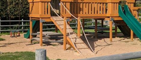 A playground with wooden structures, including a slide and climbing area, surrounded by a fence.