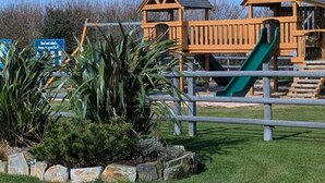 Un parque infantil con dos casitas de madera, un tobogán verde y un letrero al fondo.