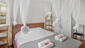 Una cama bien hecha con ropa de cama blanca y un plaid gris, una mesita de noche de madera con una lámpara, y una silla con toallas blancas.