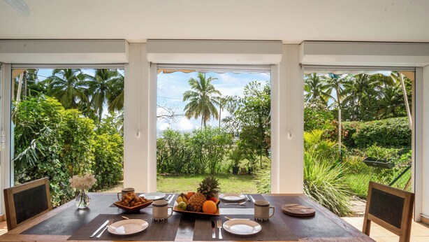 Una mesa de comedor dispuesta con platos, cubiertos y un frutero, con vistas a abundante vegetación y palmeras a través de grandes puertas de cristal.