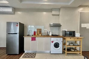 A compact kitchen with a refrigerator, washing machine, microwave, and wooden table with dishes. - Homesuite' Home at Riverson Soho (Kota Kinabalu)