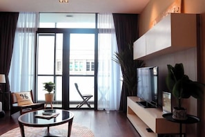 A modern living room with a glass coffee table, a flat-screen TV, a sofa, and a balcony with a view of buildings. - Homesuite' Home at Riverson Soho (Kota Kinabalu)
