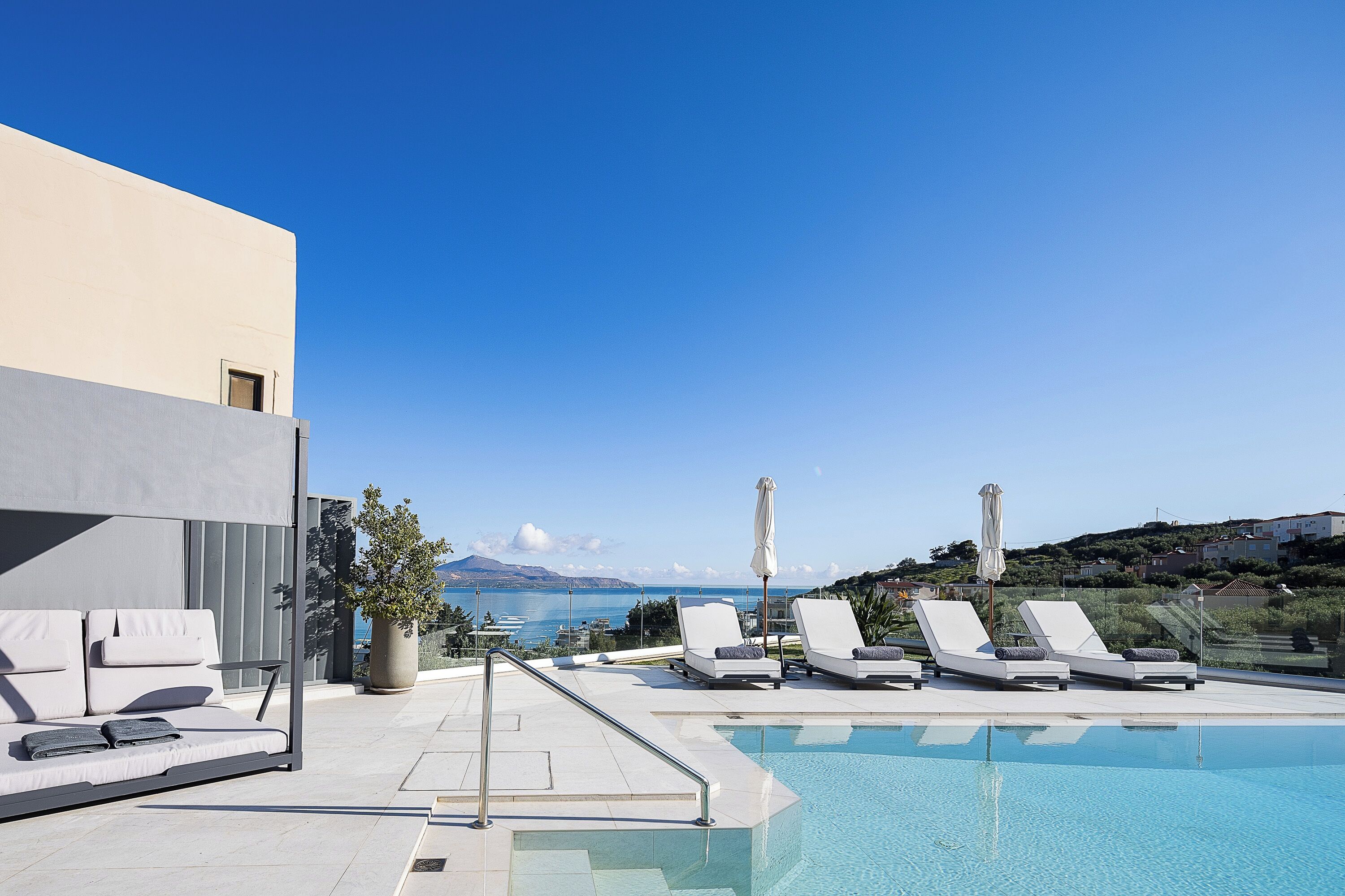 Un área de piscina en la azotea con sillones de descanso y un sofá, con vistas panorámicas del mar y las colinas.