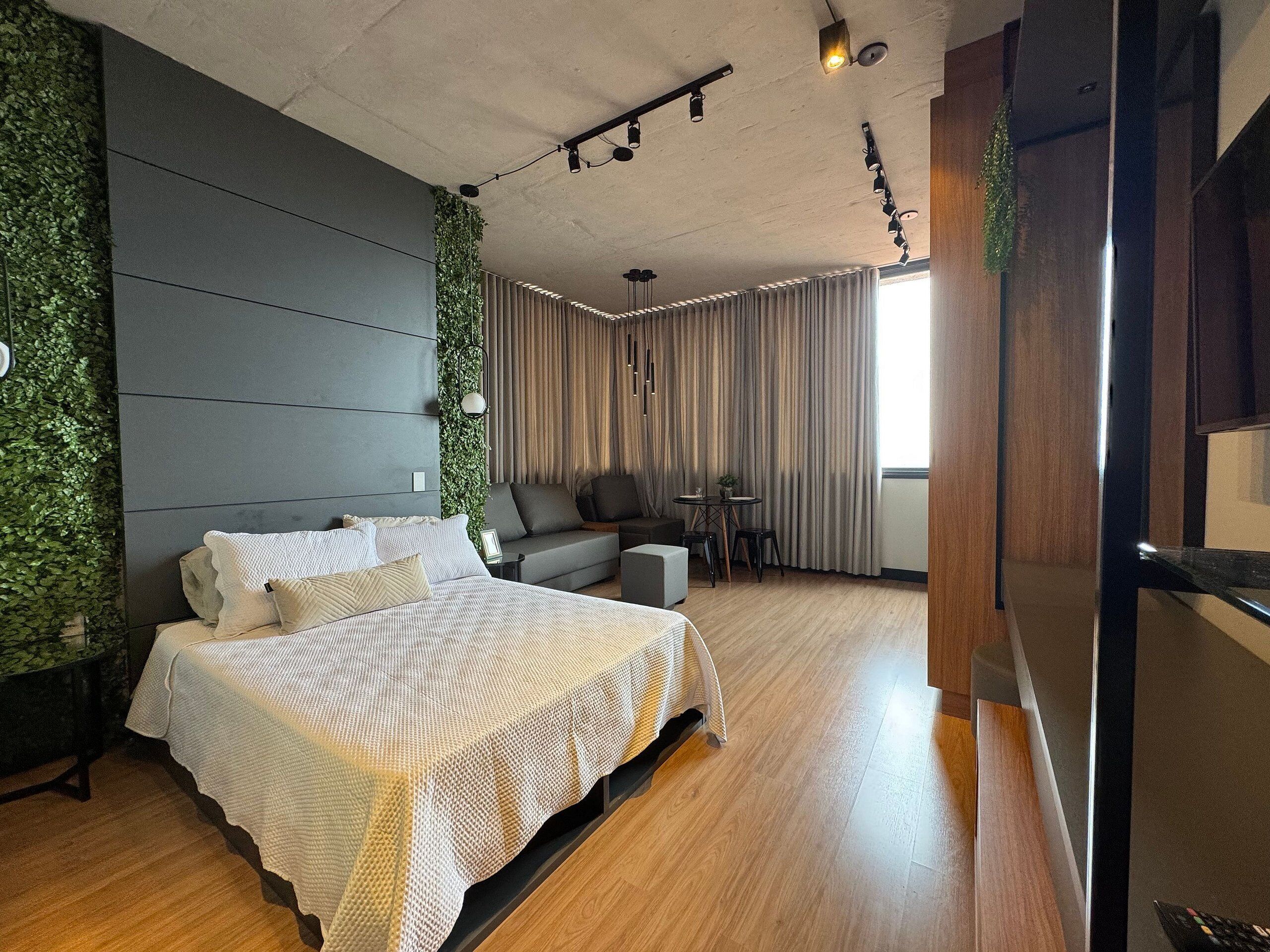 Un dormitorio moderno con una cama grande, un sofá gris, una mesita y vistas al exterior a través de una ventana.