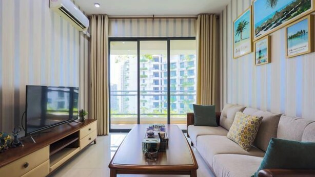 A modern living room with a flat-screen TV, a wooden coffee table, a sofa, and a large window with a view of high-rise buildings.