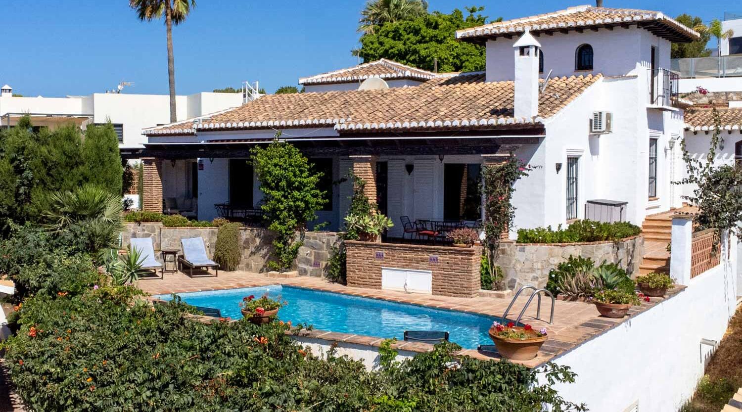 A two-story house with a swimming pool, surrounded by greenery and a clear blue sky.