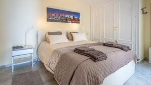 Une chambre à coucher bien rangée, avec un grand lit, une table de chevet avec une lampe, un tableau représentant un paysage urbain accroché au mur et une armoire.